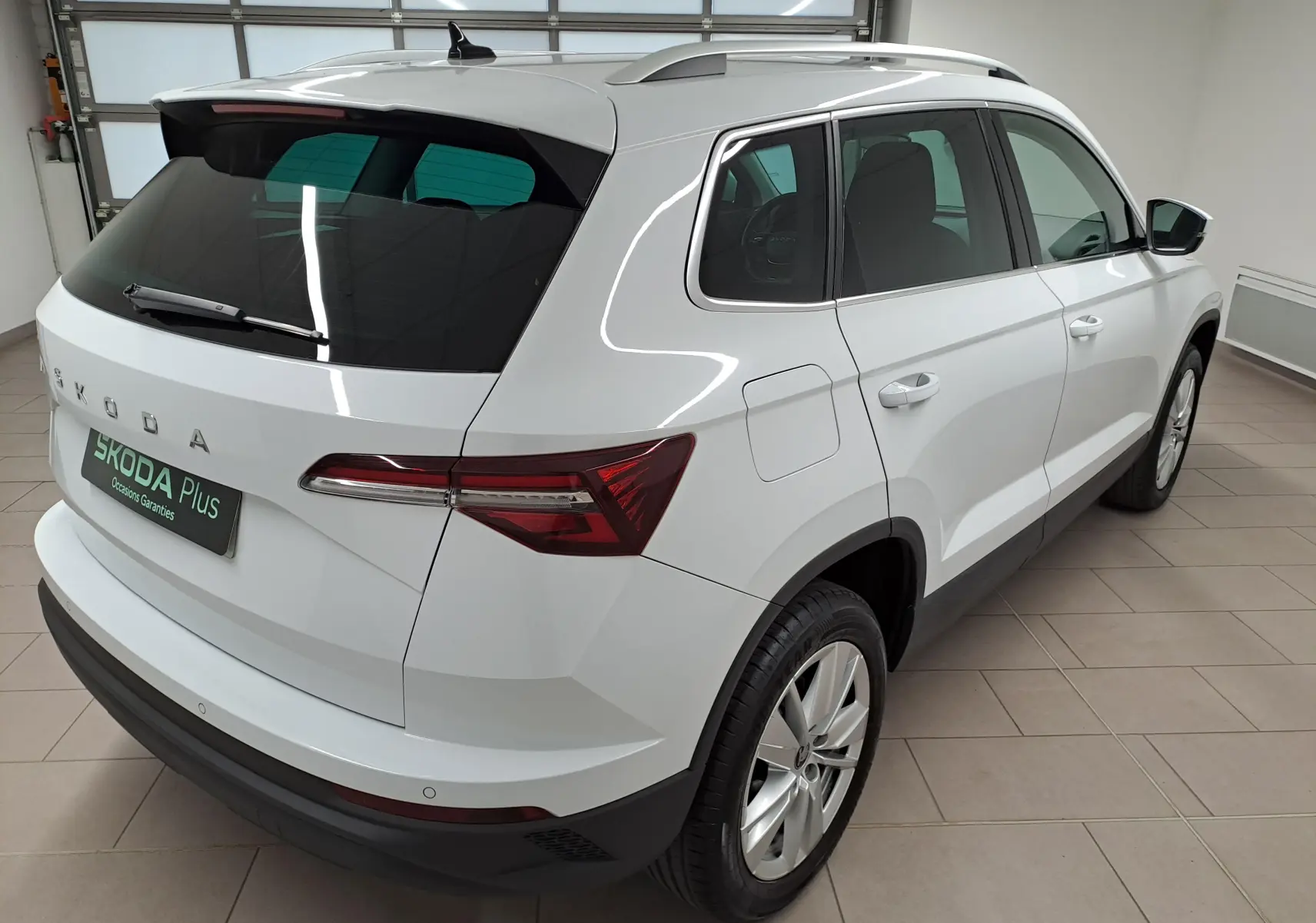 Vue 3/4 arrière droite d'un Skoda Karoq blanc lune 2025 avec vitres teintées et jantes alliage en intérieur showroom.