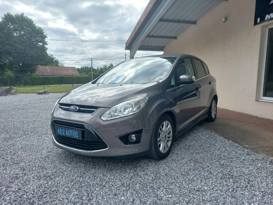 Ford C-MAX gris en 3/4 avant droit, garé sur gravier devant un bâtiment avec ciel nuageux.