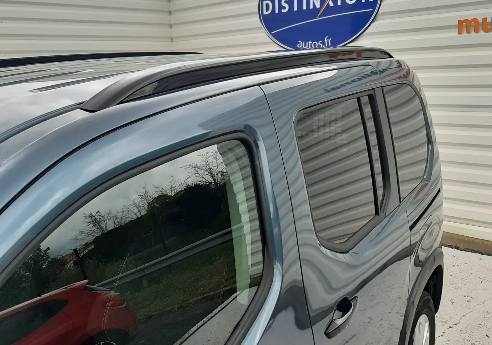 Vue 3/4 arrière côté droit du Peugeot Rifter bleu Kiama avec barres de toit noir mat et vitres arrière surteintées.