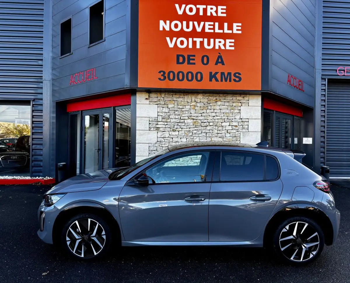 Profil côté gauche d'une Peugeot 208 Hybrid 136 eDCS6 GT gris foncé stationnée devant un bâtiment avec panneau orange.