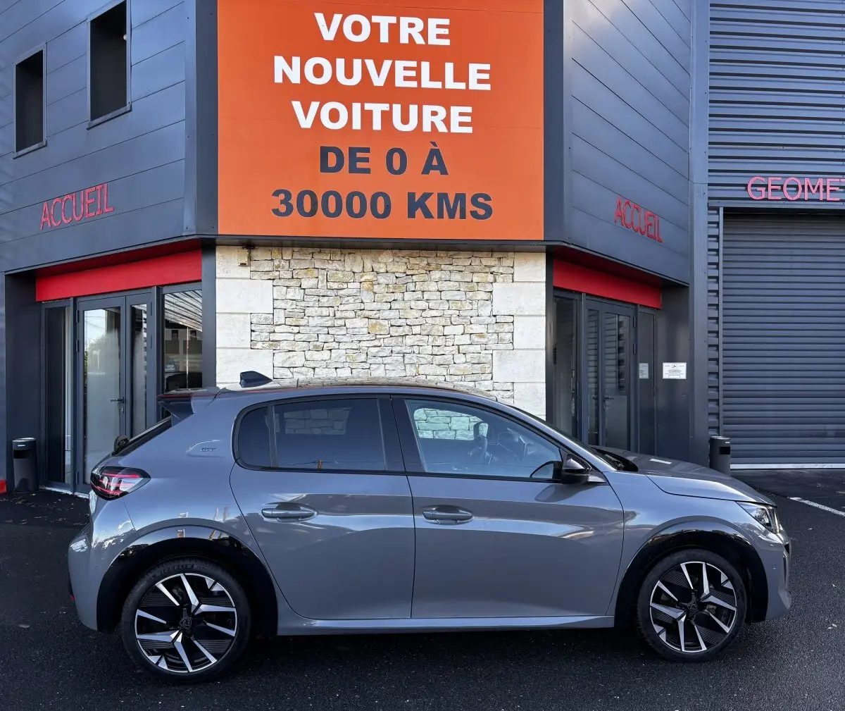 Profil côté droit d'une Peugeot 208 Hybrid 136 eDCS6 GT gris foncé garée devant un bâtiment commercial.