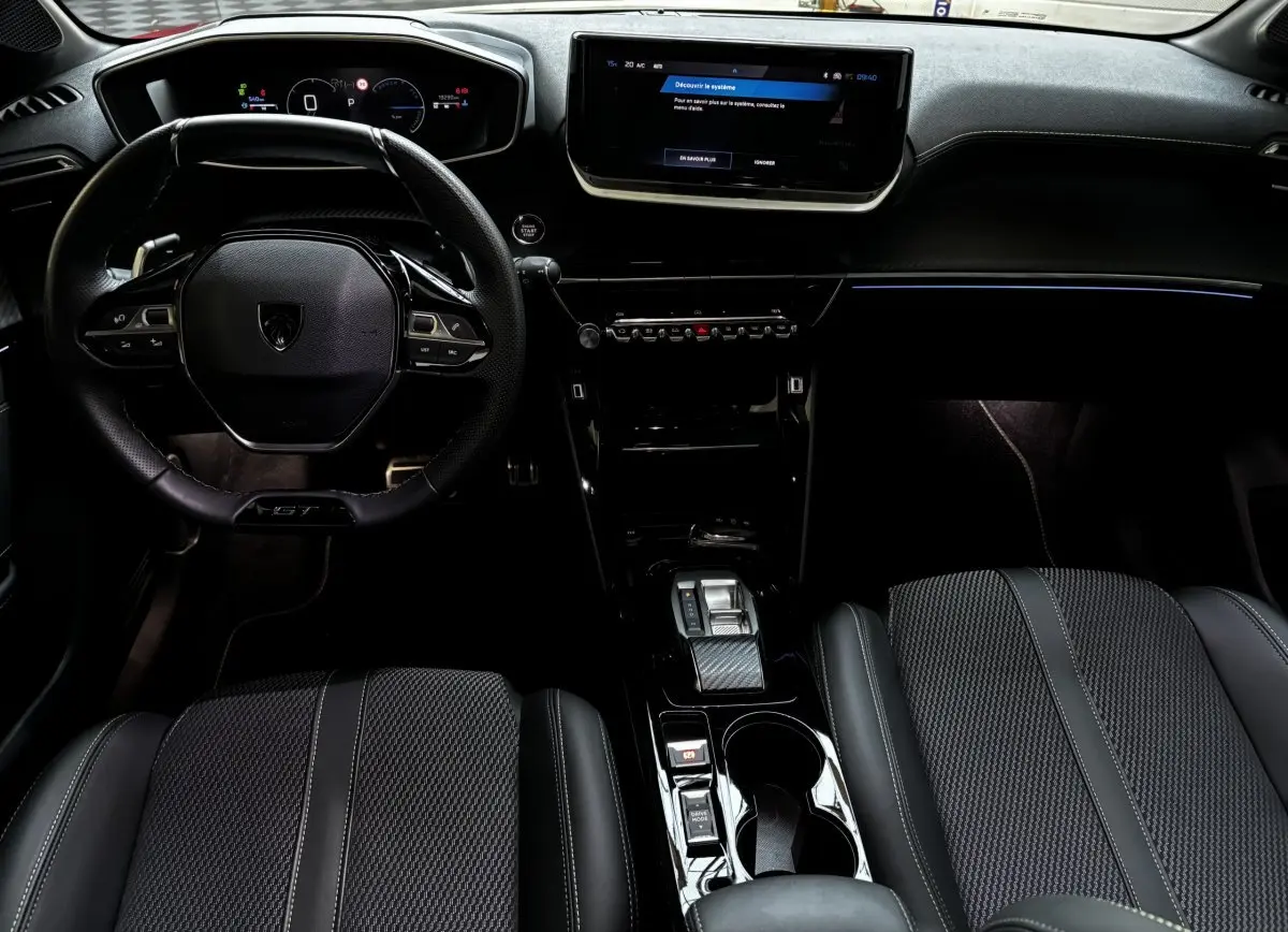 Intérieur Peugeot 208 Hybrid 136 GT vu de face, volant cuir, écran tactile central et sièges noirs texturés.