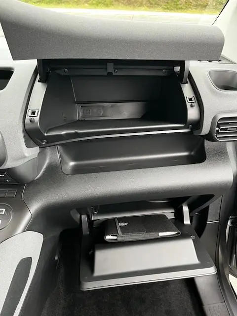 Vue intérieure du tableau de bord du Peugeot Rifter 2024 gris foncé, avec boîte à gants ouverte et rangement supérieur visible.
