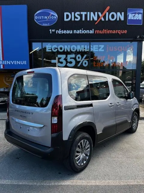 Vue 3/4 arrière droite du Peugeot Rifter gris foncé 2024 avec vitres opaques et rails de toit, stationné devant un concessionnaire.