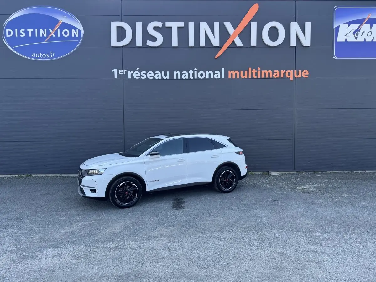 Vue de profil droit d'un DS7 Crossback blanc stationné devant un mur gris avec logo Distinxion.