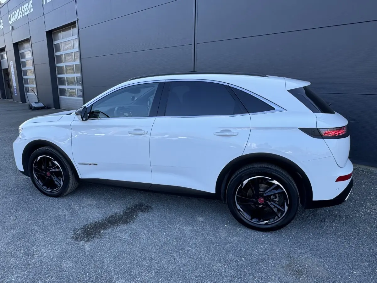 Vue de profil côté gauche d'un DS7 Crossback blanc 2020 avec jantes noires et vitres teintées, stationné devant un bâtiment.
