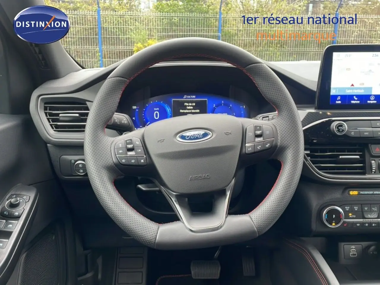 Vue intérieure centrée sur le volant perforé avec coutures rouges du Ford Kuga 2024, tableau de bord digital et écran tactile visible.