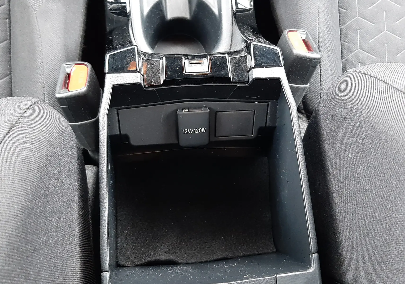 Vue plongeante sur la console centrale noire de la Toyota Corolla blanche 122 CH Dynamic, avec prise 12V visible entre les sièges en tissu gris.