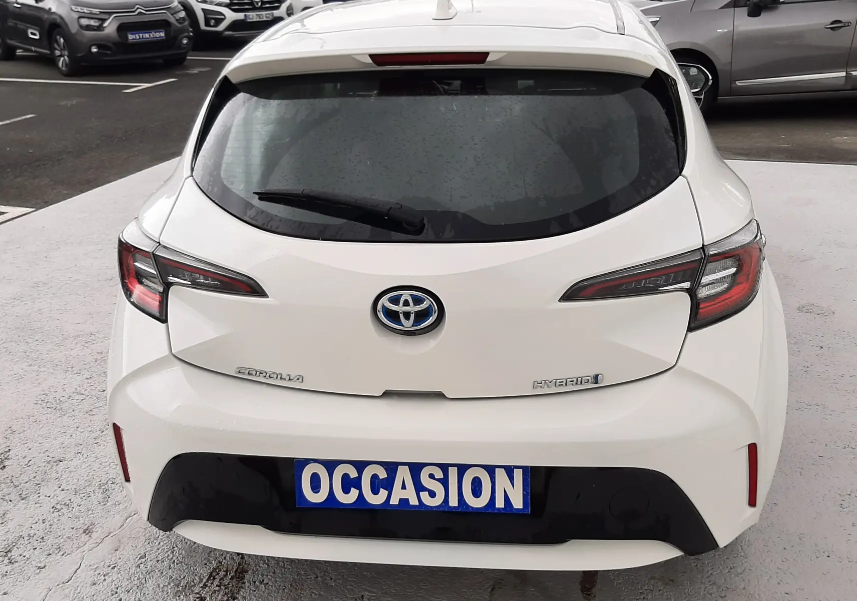 Vue arrière d'une Toyota Corolla blanche hybride 2022 avec plaque "OCCASION" sur un parking de voitures.