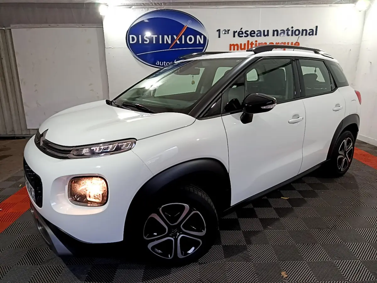 Vue 3/4 avant droite d'un Citroën C3 Aircross blanc avec phares allumés et barres de toit noires en intérieur showroom.