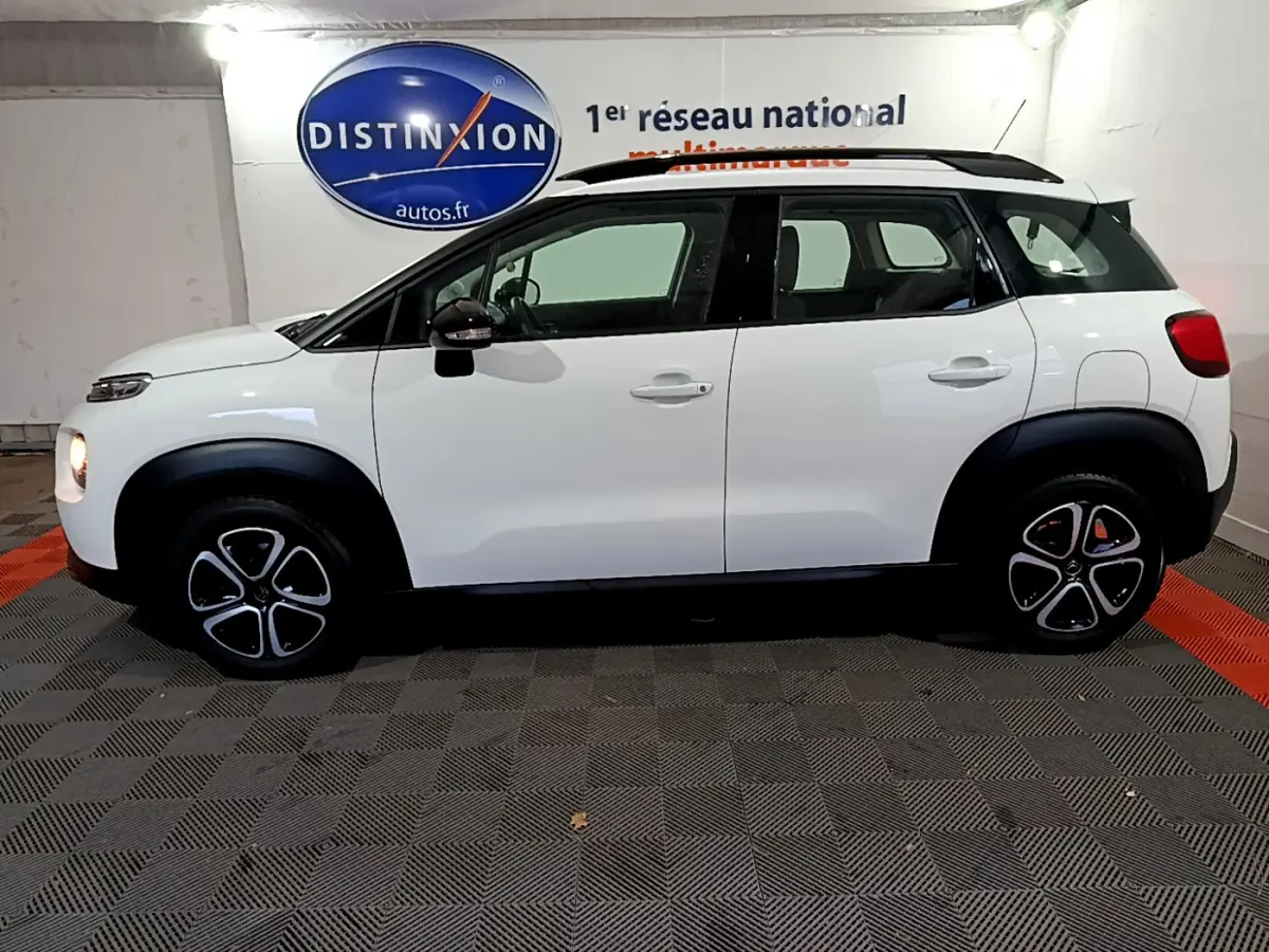 Profil côté gauche d'un Citroën C3 Aircross blanc avec toit noir et jantes au design distinctif en intérieur showroom.