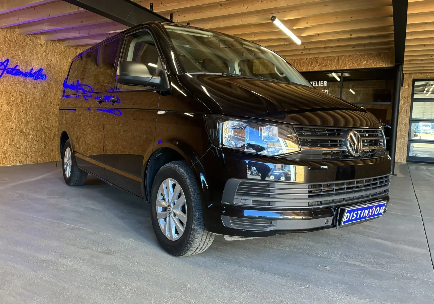 Volkswagen Multivan T6 noir vu en 3/4 avant droit dans un garage avec mur en bois et néon bleu.
