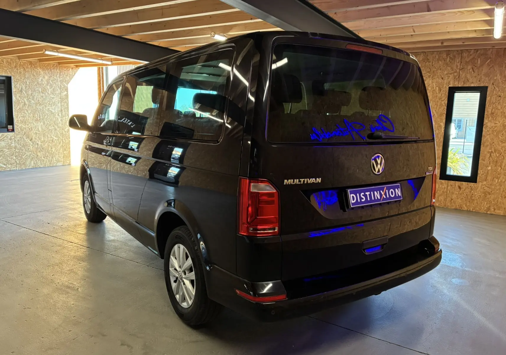 Volkswagen Multivan T6 noir vu en 3/4 arrière droit dans un garage avec reflets bleutés sur la carrosserie.