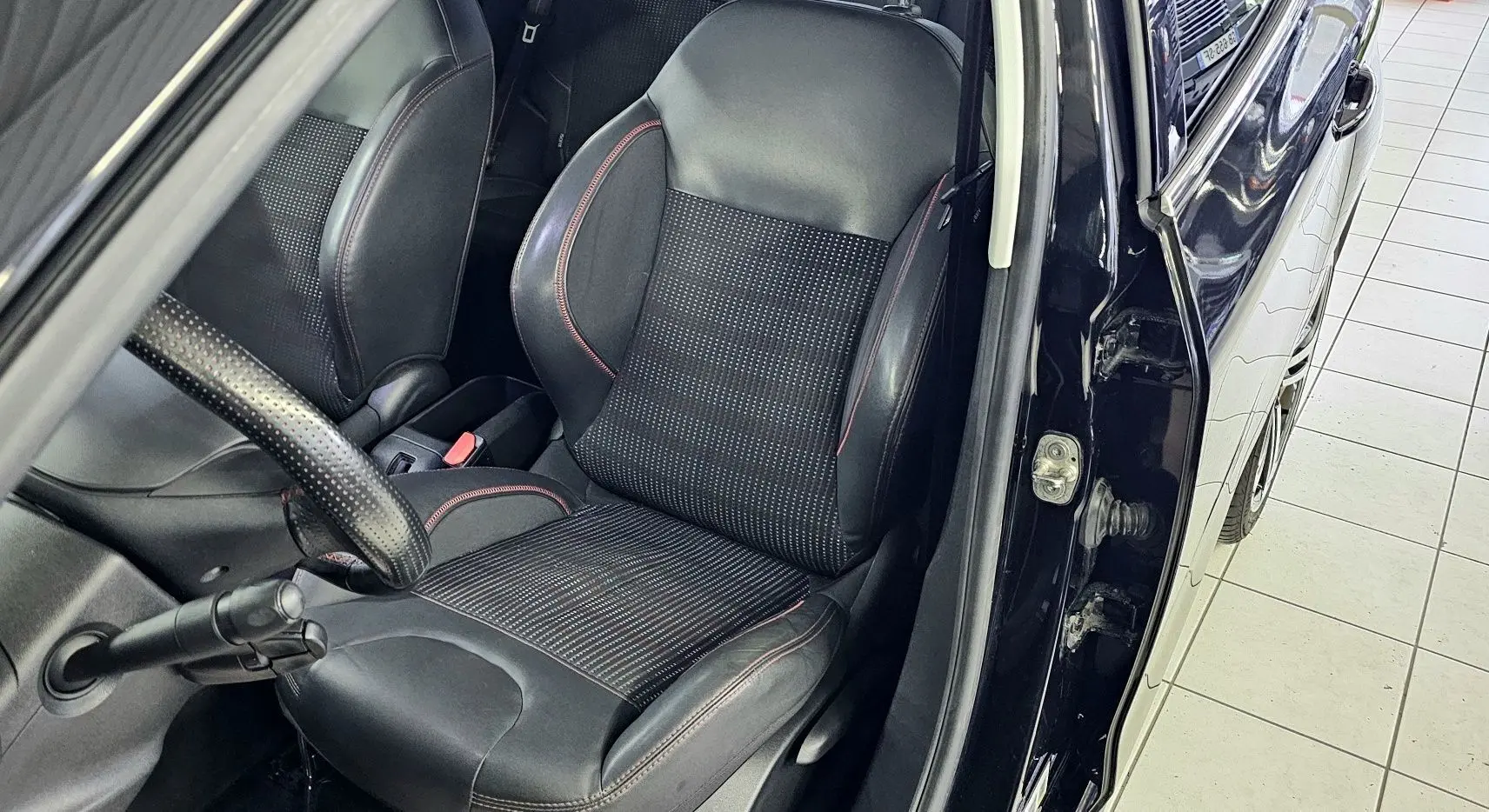 Vue intérieure côté conducteur d'une Peugeot 208 GT-LINE noire de 2015, siège en cuir et tissu avec surpiqûres rouges.
