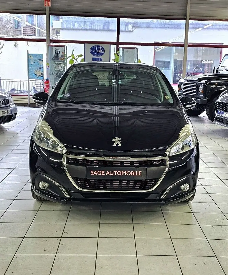 Vue de face d'une Peugeot 208 GT-LINE noire, avec calandre chromée et phares allongés en showroom.