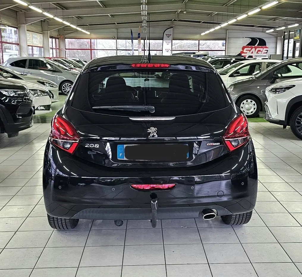 Vue arrière d'une Peugeot 208 GT-LINE noire stationnée en intérieur avec feux arrière allumés et attelage visible.