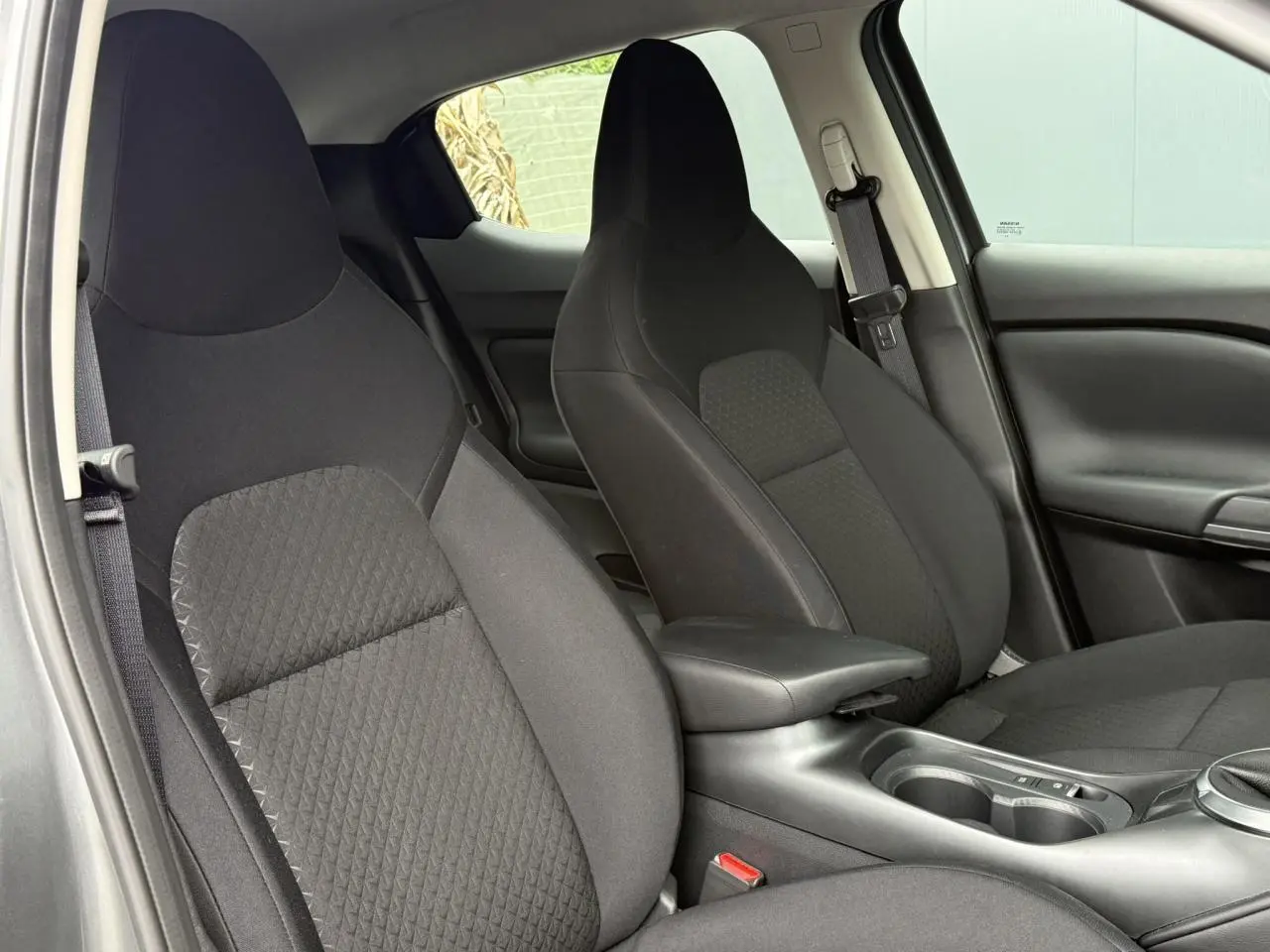 Vue intérieure rapprochée des sièges avant noirs du Nissan Juke 2025, avec accoudoir central et ceintures visibles.