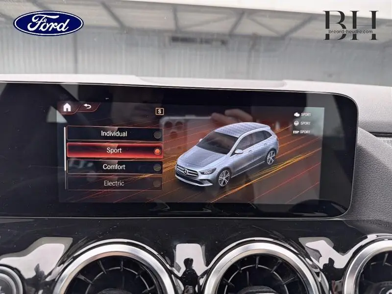 Écran tactile intérieur montrant une Mercedes Classe B grise en vue 3/4 avant avec mode Sport sélectionné.