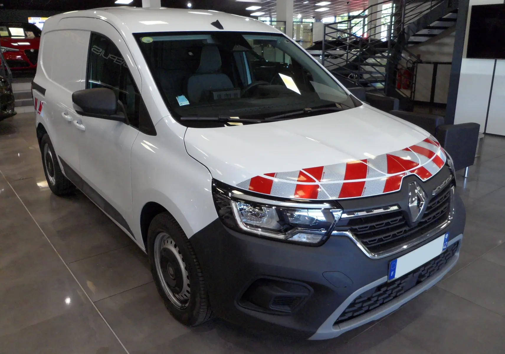 Renault Kangoo Van blanc minéral en 3/4 avant droit avec bandes réfléchissantes rouges sur le capot, en intérieur showroom.