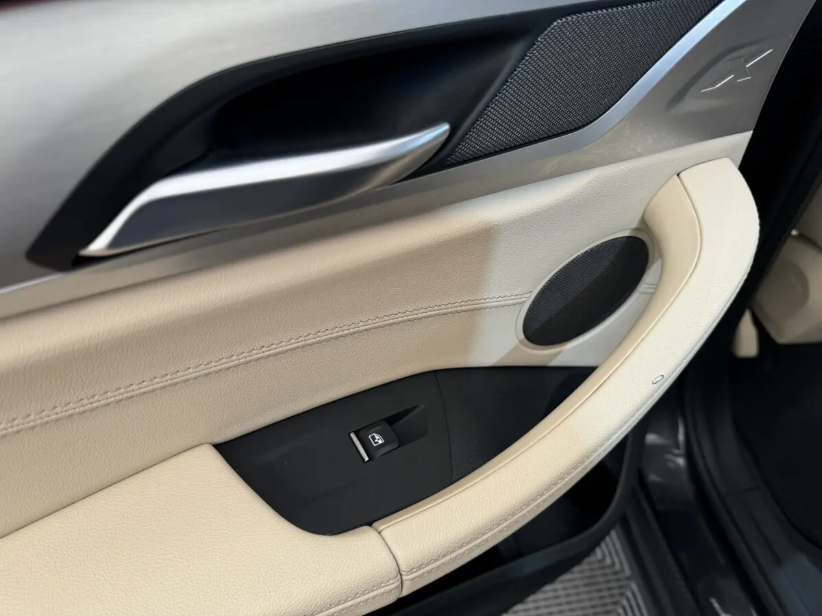 Poignée de porte intérieure beige côté conducteur du BMW X3 xDrive30d 2018 avec bouton de lève-vitre et haut-parleur intégré.