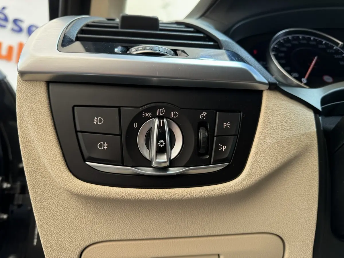Gros plan sur le panneau de commande des phares du BMW X3 gris foncé, intérieur beige visible autour.