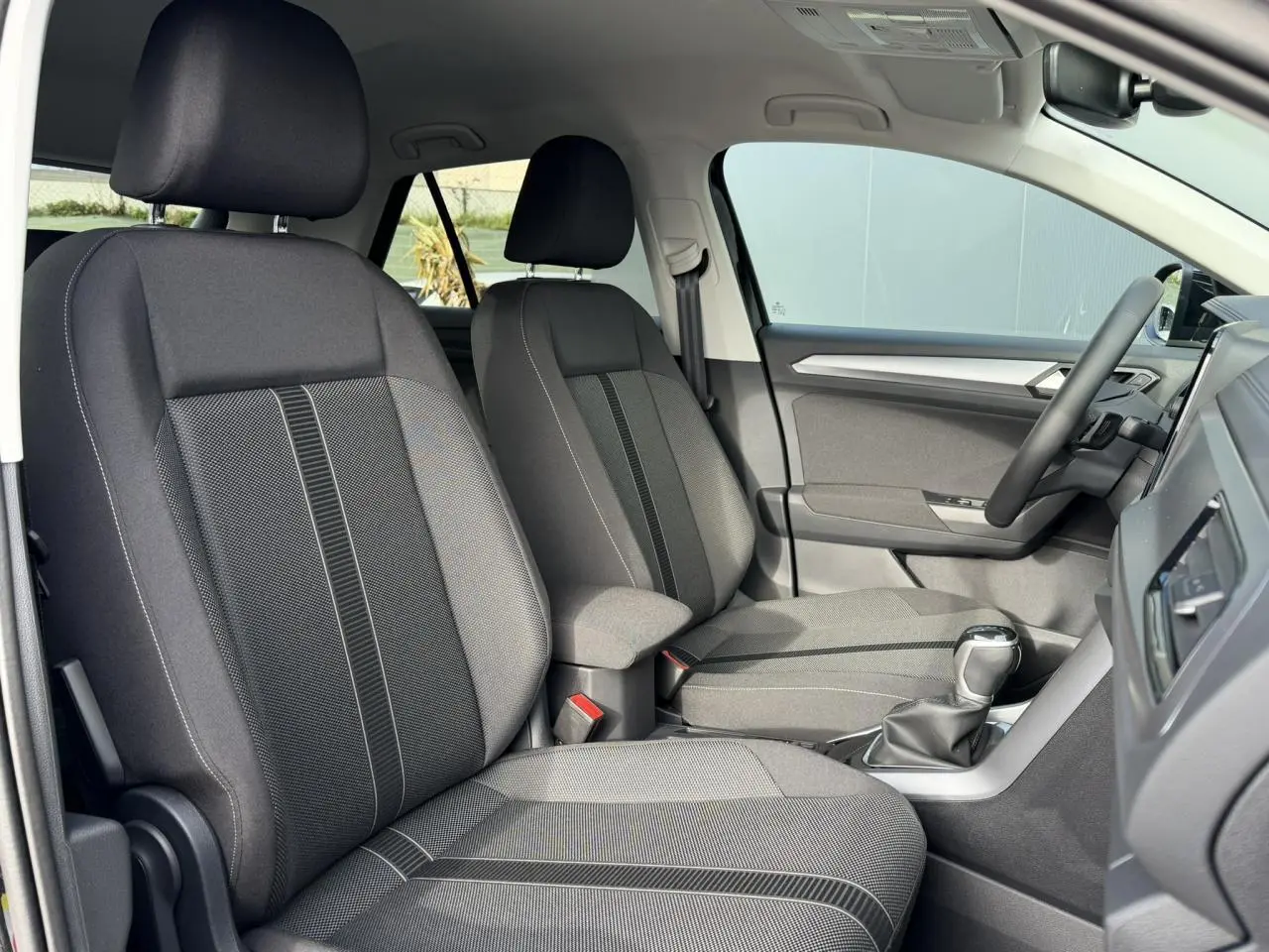 Vue intérieure côté gauche du Volkswagen T-Roc 1.5 TSI 2025, sièges tissu gris avec accoudoir central.