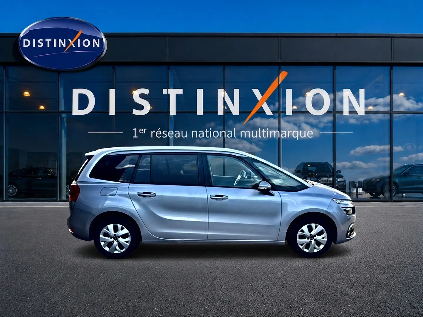 Profil côté gauche d'un Citroën Grand C4 Spacetourer gris alu 2021 garé devant une vitrine avec logo Distinxion.
