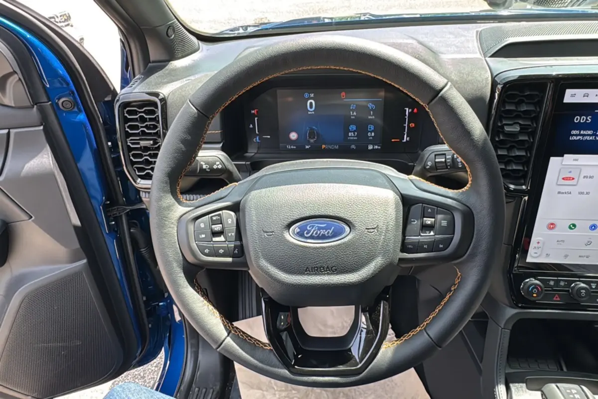 Vue intérieure centrée sur le volant cuir noir avec surpiqûres marron du Ford Ranger Wildtrak X bleu lightning, tableau de bord digital visible.