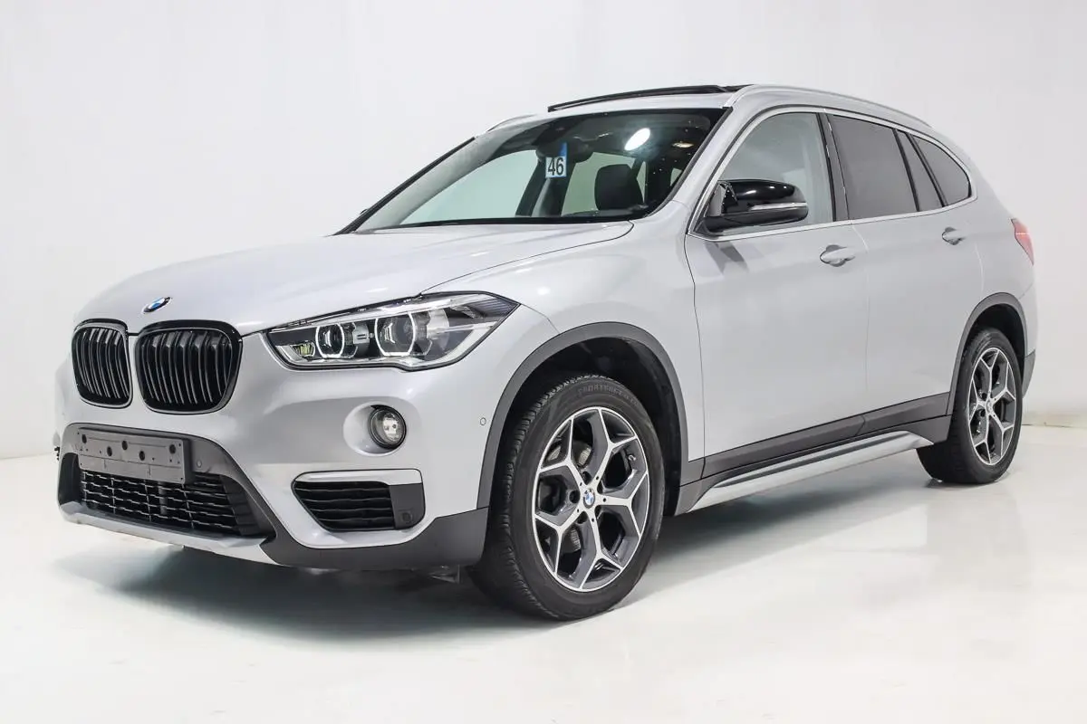 BMW X1 sDrive 18D argent métallisé en 3/4 avant droit avec toit ouvrant et attelage visible.