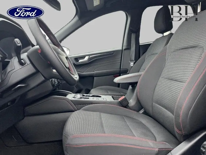 Vue intérieure côté conducteur du Ford Kuga 2023 avec sièges sport noirs et coutures rouges, volant cuir multifonction.