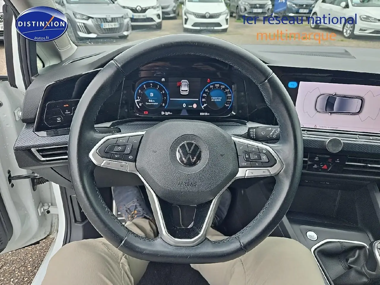 Vue intérieure centrée sur le volant cuir noir d'une Volkswagen Golf blanche, avec tableau de bord numérique et écran tactile visible.
