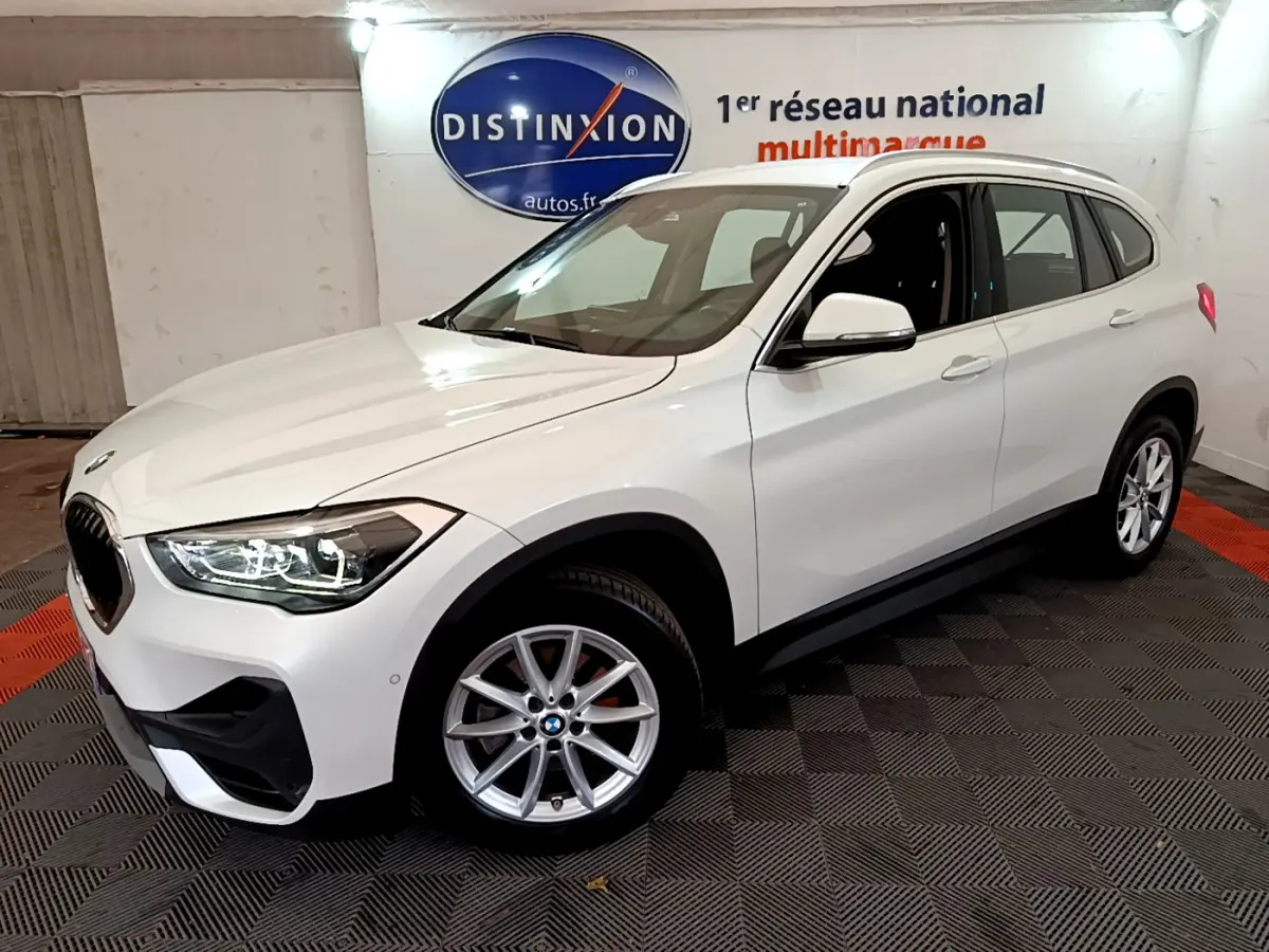 BMW X1 sDrive18i blanc vue 3/4 avant droit, phares LED allumés et jantes alliage visibles.