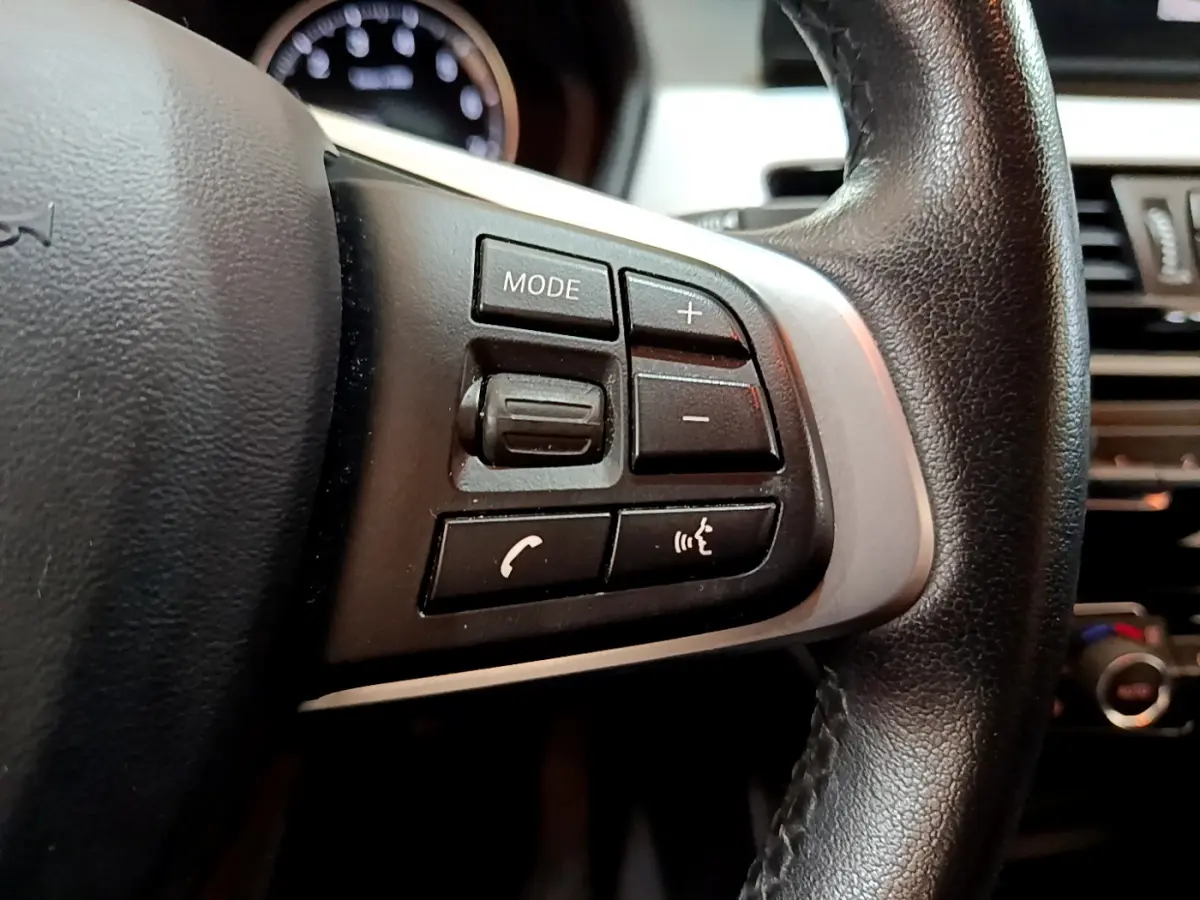 Gros plan sur les commandes gauche du volant cuir noir du BMW X1 sDrive18i Lounge 2019, avec boutons multifonctions.