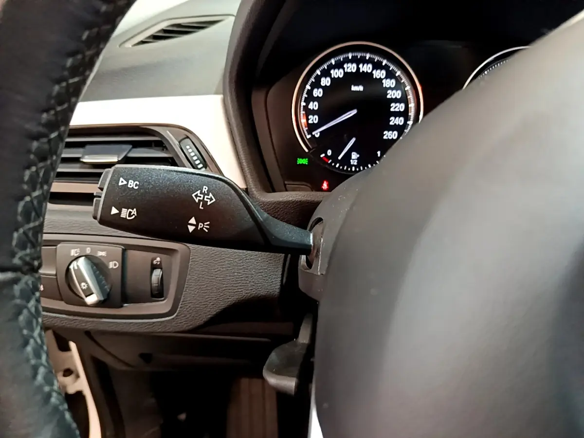 Gros plan sur le volant cuir et le compteur noir du tableau de bord d'une BMW X1 sDrive18i Lounge blanche, vue côté conducteur.