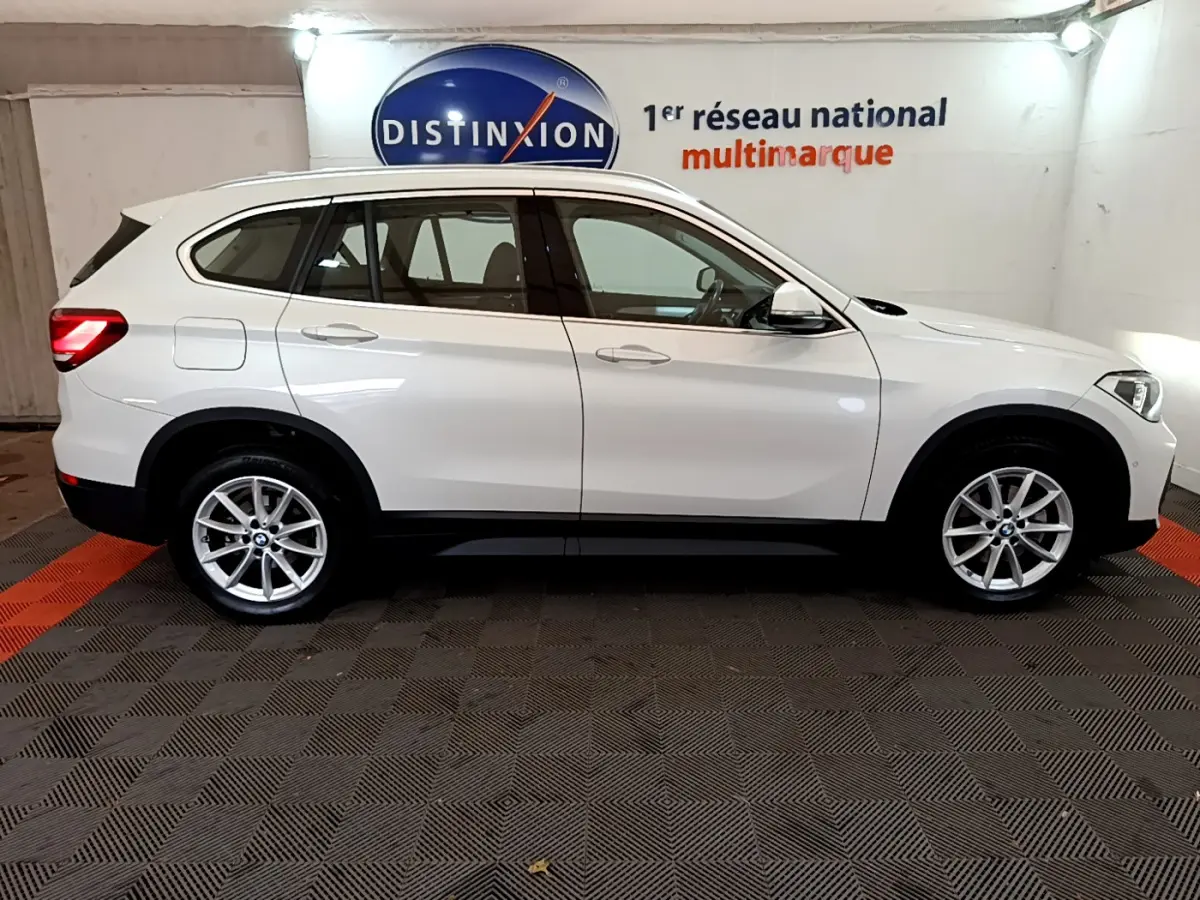 BMW X1 sDrive18i blanc vue profil côté gauche dans un showroom avec jantes alliage et barres de toit noires.