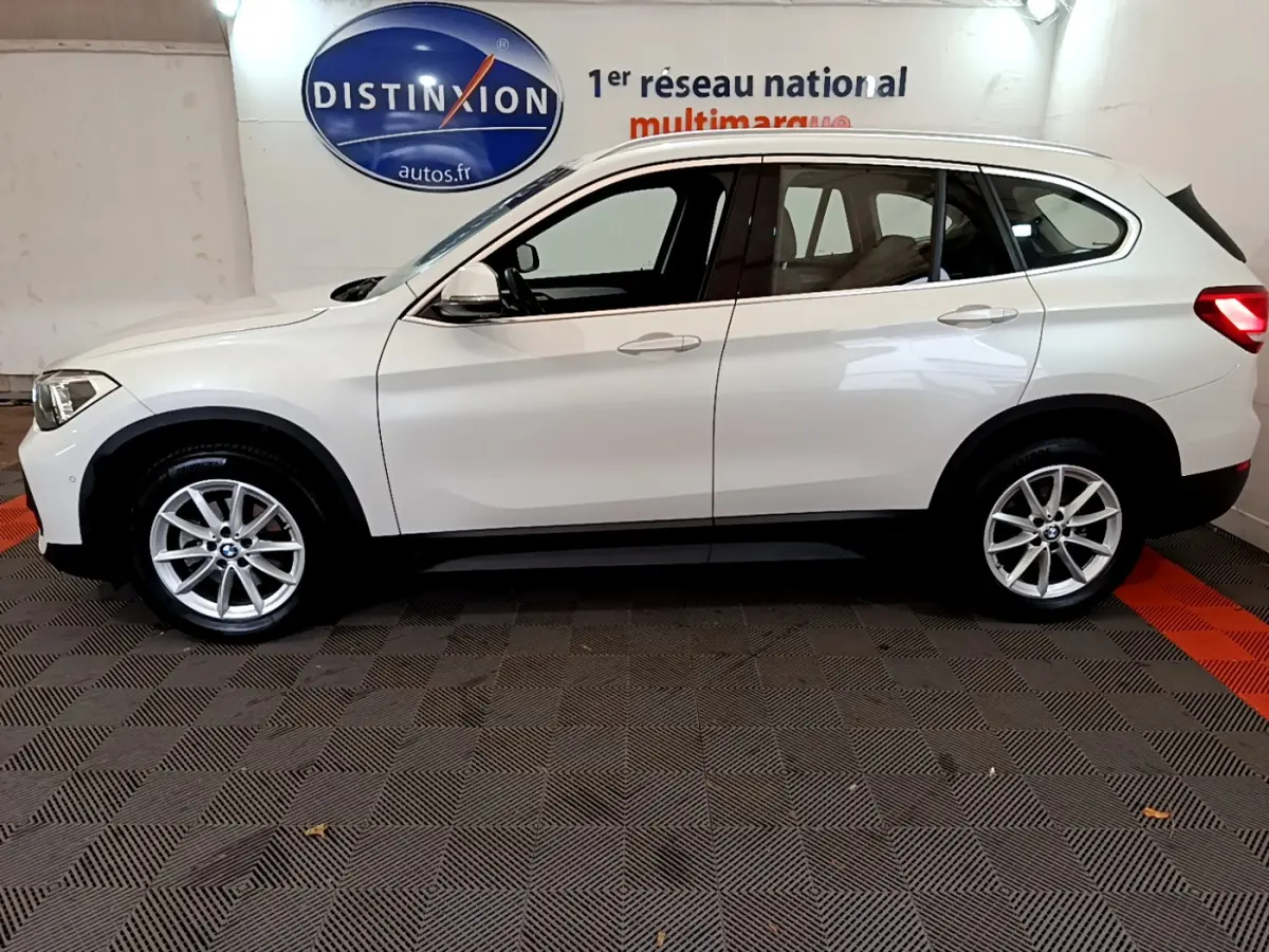 Vue de profil côté gauche d'un BMW X1 sDrive18i Lounge blanc, avec jantes alliage et rails de toit noirs.