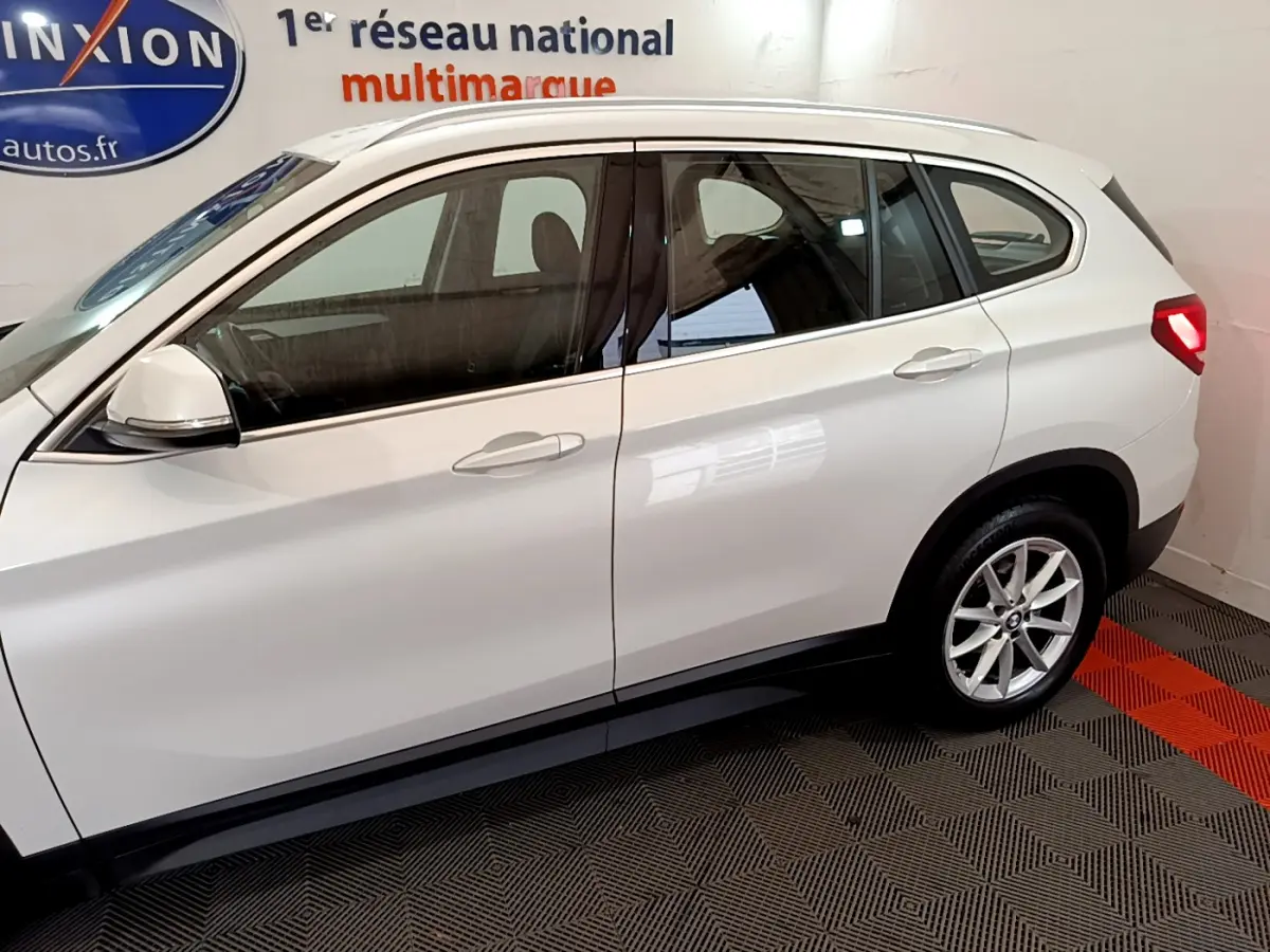 BMW X1 sDrive18i blanc vue de profil côté gauche en intérieur avec jantes alliage et rétroviseurs blancs.