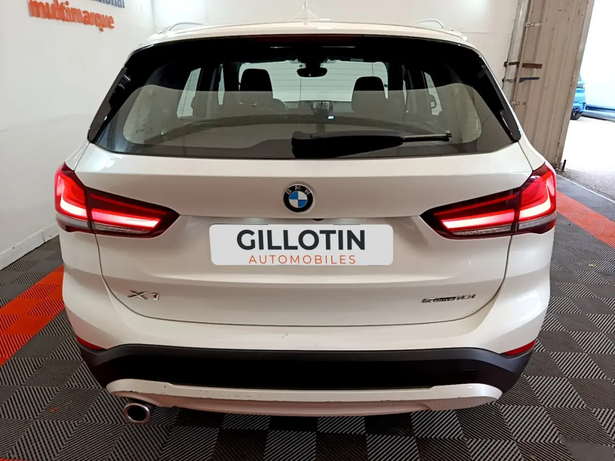 Vue arrière d'un BMW X1 sDrive18i blanc avec feux LED allumés, dans un garage au sol carrelé noir et rouge.