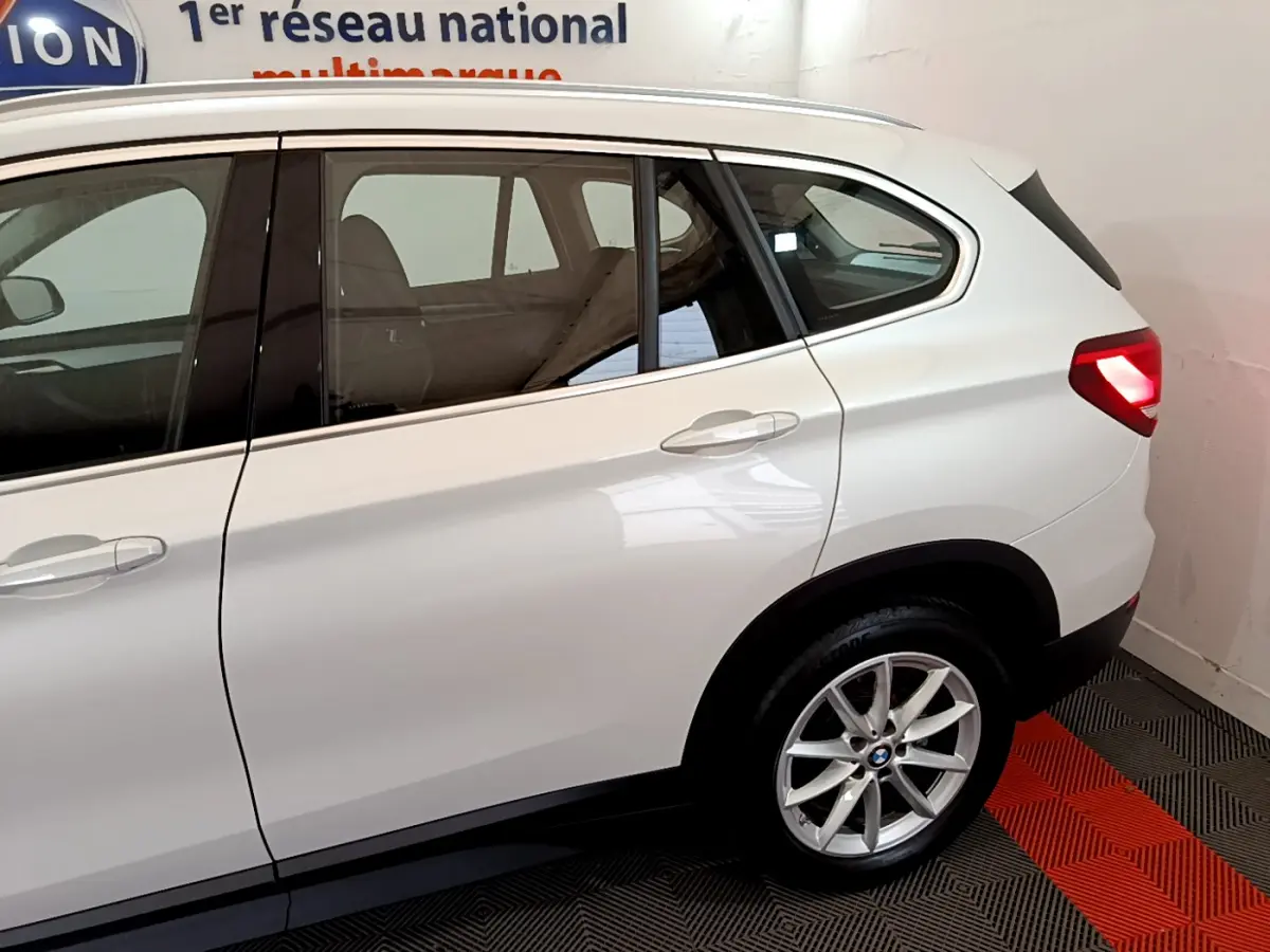 Vue côté droit arrière d'un BMW X1 sDrive18i Lounge blanc avec feux arrière allumés dans un showroom.