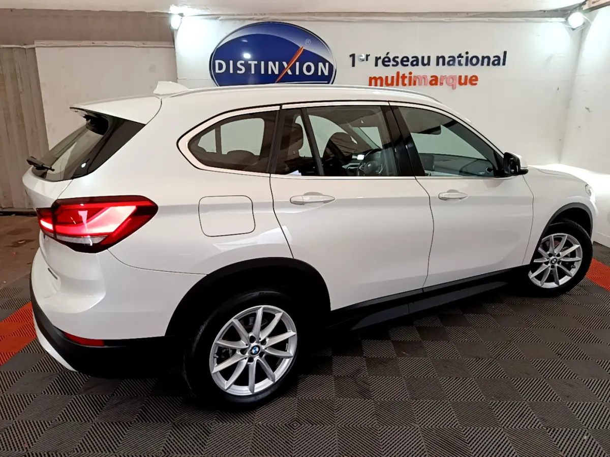 BMW X1 sDrive18i blanc en vue profil droit, feux arrière allumés, roues alliage et toit avec rails noirs.