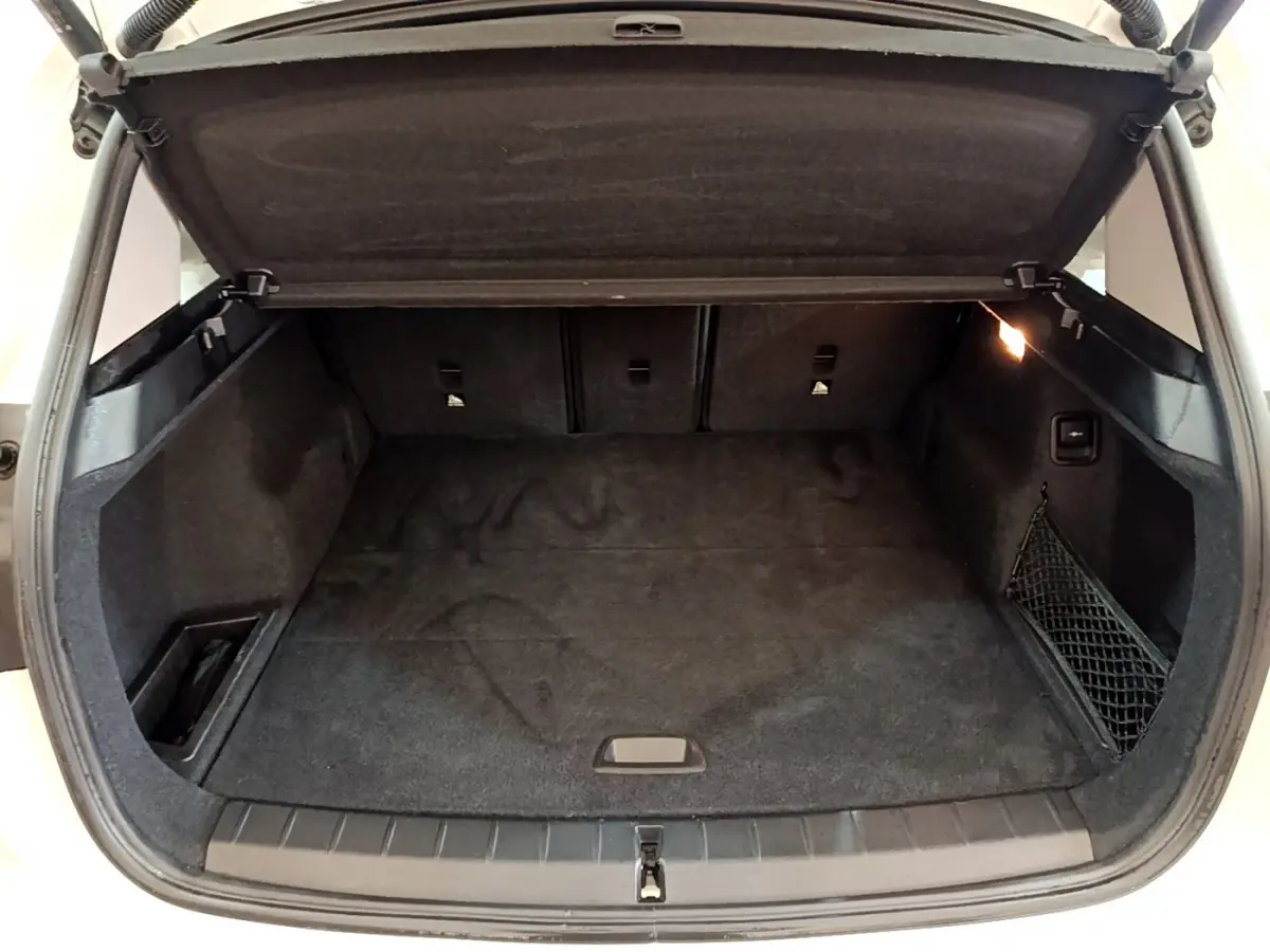 Coffre ouvert de BMW X1 sDrive18i blanc, vue arrière, espace de chargement avec tapis noir et filet latéral.
