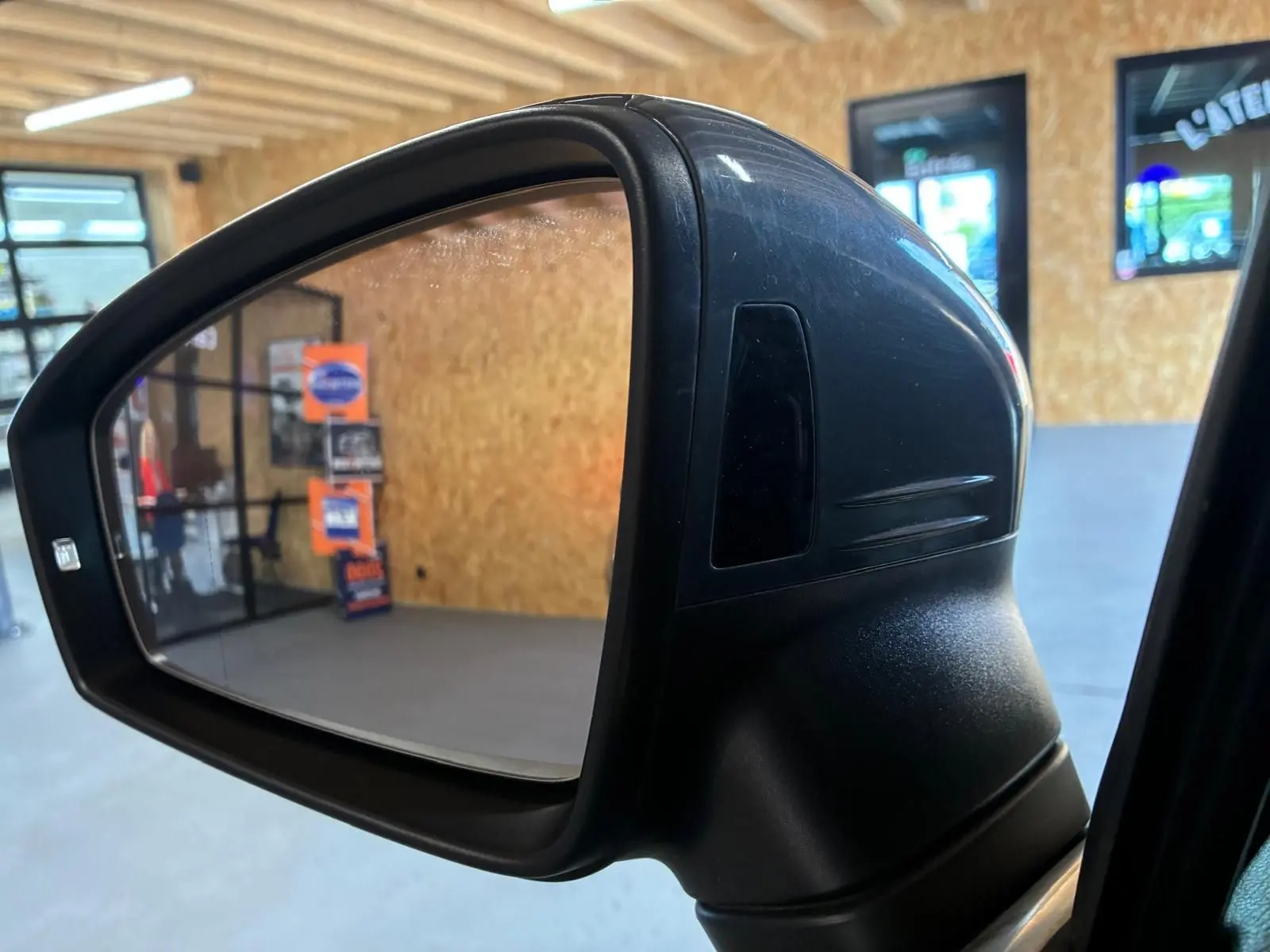 Gros plan sur le rétroviseur droit noir du Volkswagen Tiguan gris, intérieur d'atelier visible en reflet.