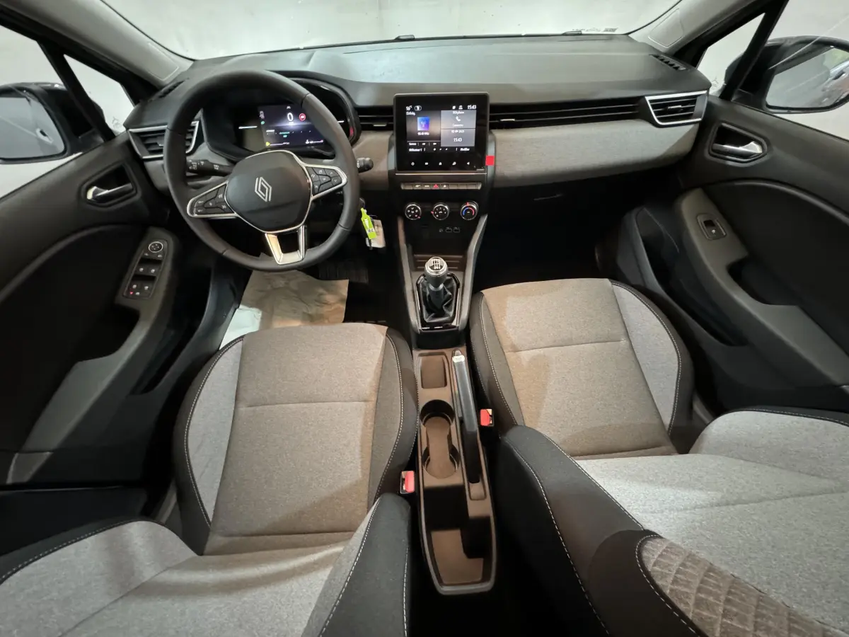 Vue intérieure avant de la Renault Clio gris clair 2025, avec volant multifonctions, écran tactile central et sièges tissu bicolore.