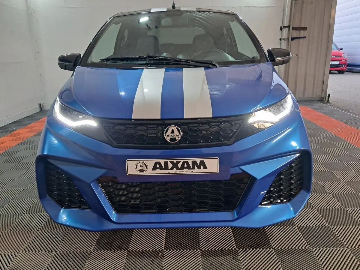 Vue frontale d'une AIXAM AMBITION 2025 bleu avec bandes blanches sur le capot et phares allumés dans un garage.