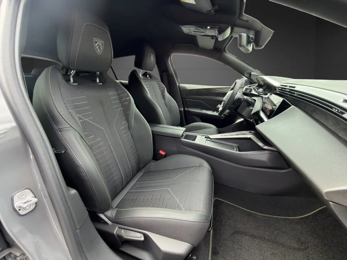 Vue intérieure côté conducteur de la Peugeot 308 GT 2026 gris clair, sièges noirs avec logo lion brodé et tableau de bord moderne.
