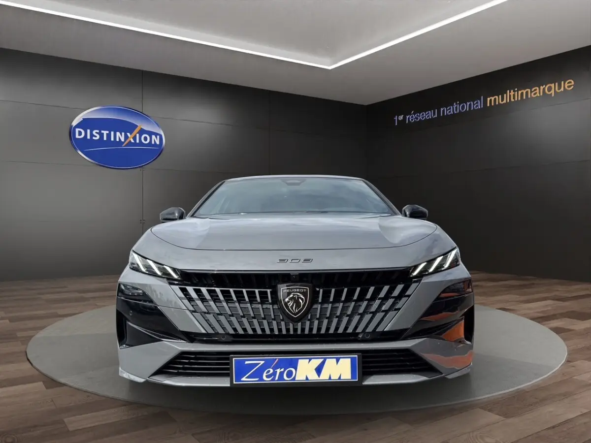 Vue de face d'une Peugeot 308 gris clair avec calandre noire distinctive et phares LED fins en intérieur showroom.