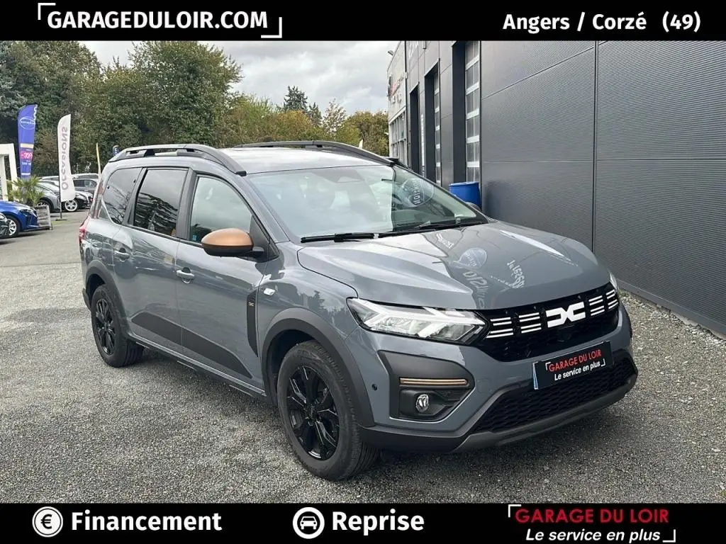 Vue 3/4 avant d’un Dacia Jogger Extreme+ Hybrid 140 gris avec rétroviseurs bronze et jantes noires, stationné devant un bâtiment.