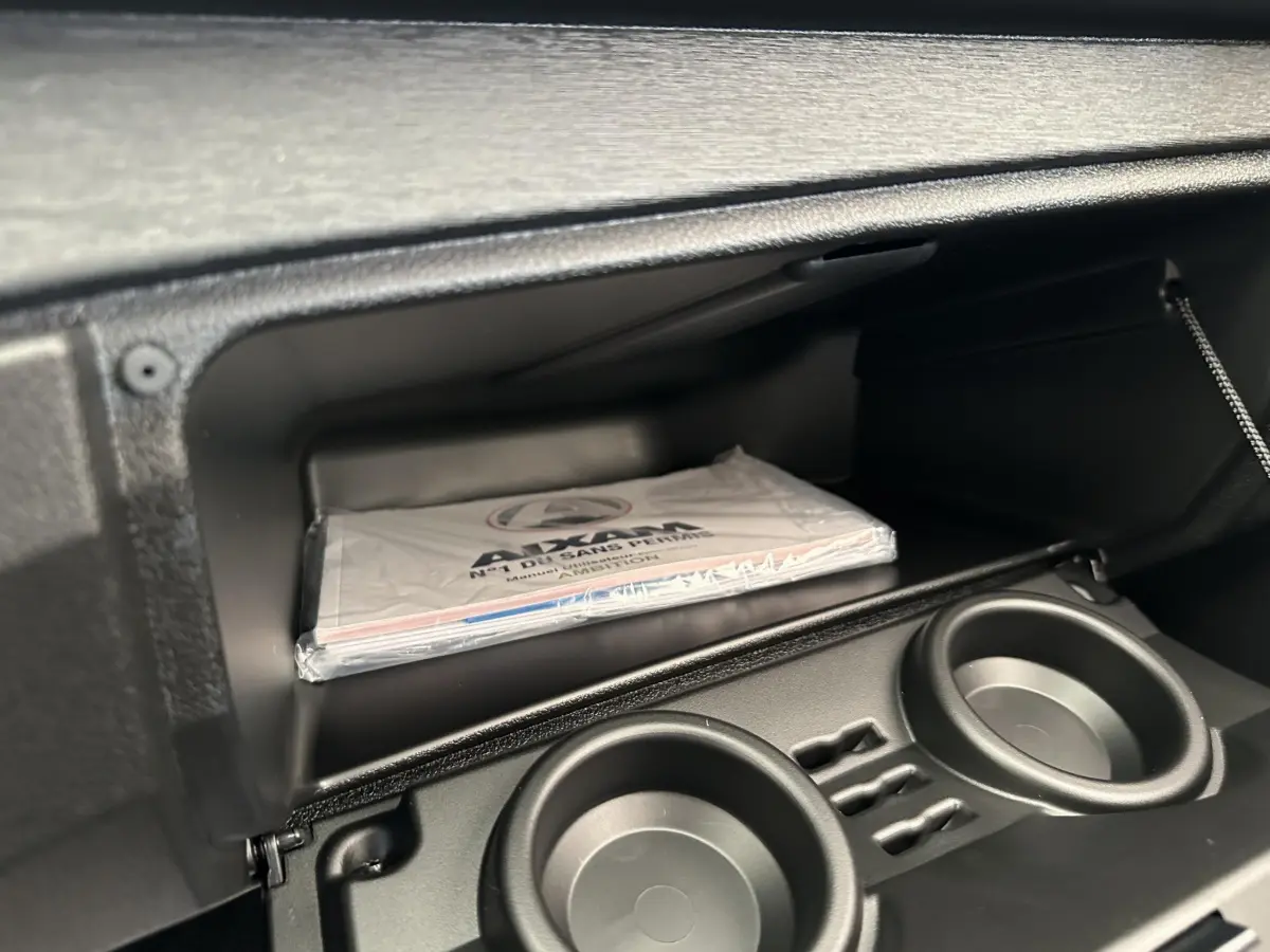 Gant de boîte à gants ouvert avec manuel AIXAM AMBITION 0.5 CITY PACK 2025 et porte-gobelets intégrés.