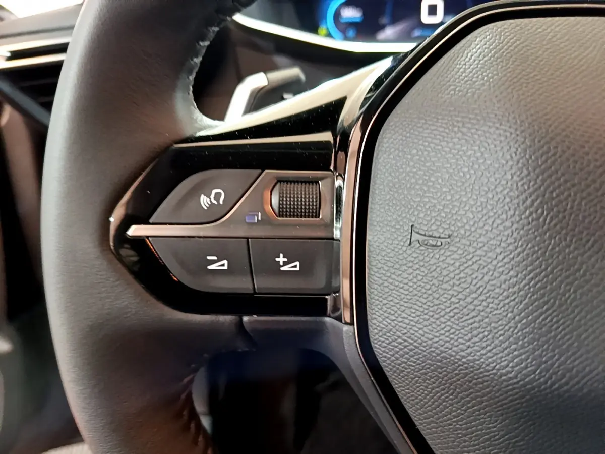 Gros plan sur le volant cuir noir de la Peugeot 208 2022, avec commandes audio et tableau de bord numérique en arrière-plan.
