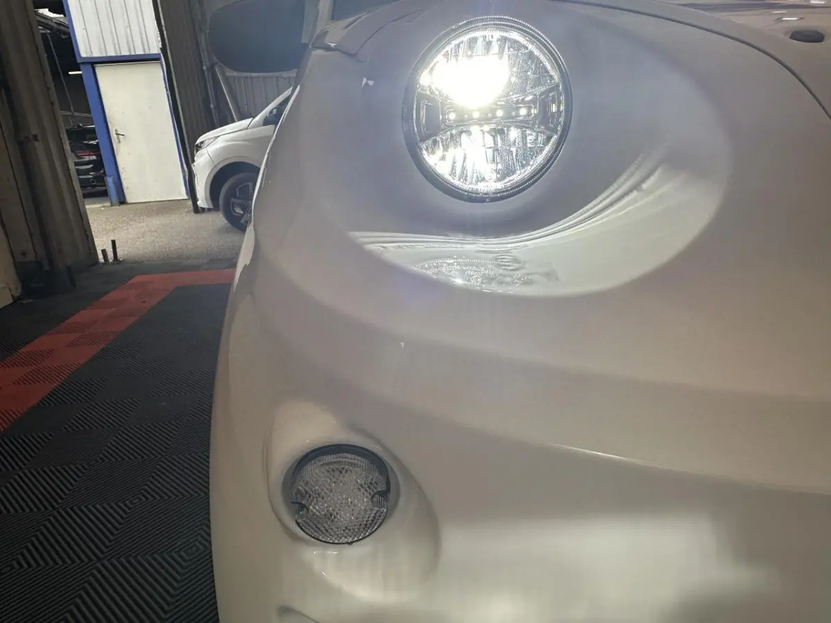 Gros plan sur le phare avant droit allumé d'une Aixam Minauto E Access blanche, avec feu antibrouillard visible.